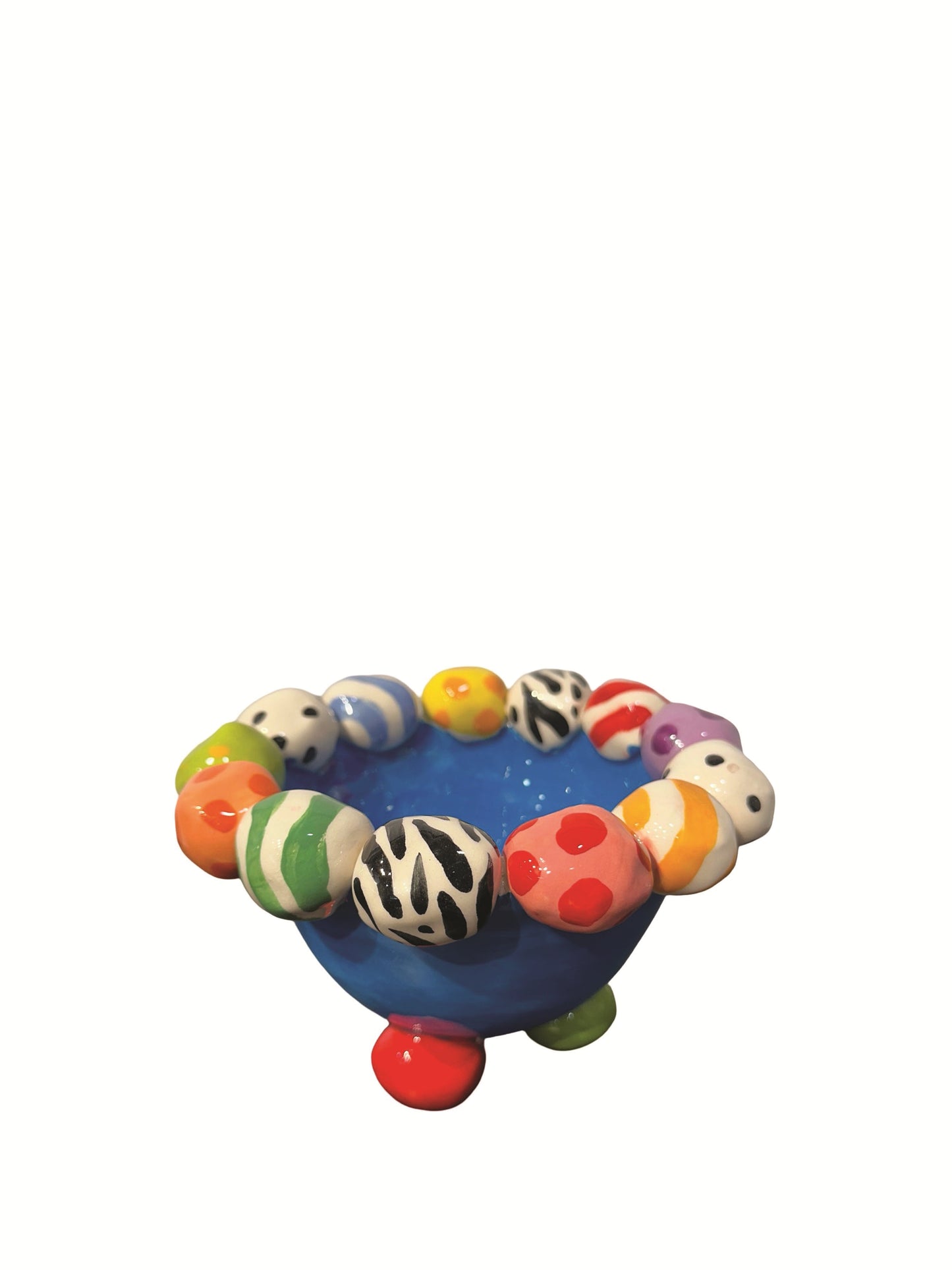 This handmade ceramic footed bowl by Mary Rose Young features a vivid blue glaze and playful hand-painted orbs in stripes, polka dots, and animal prints around its rim. It rests on sculpted red, green, and yellow feet, making it a striking decorative piece suitable for serving or display. Dimensions are approximately 6.5 inches in width and depth, and 4 inches high. Crafted from ceramic, this unique item combines functional art with bold, colorful design.