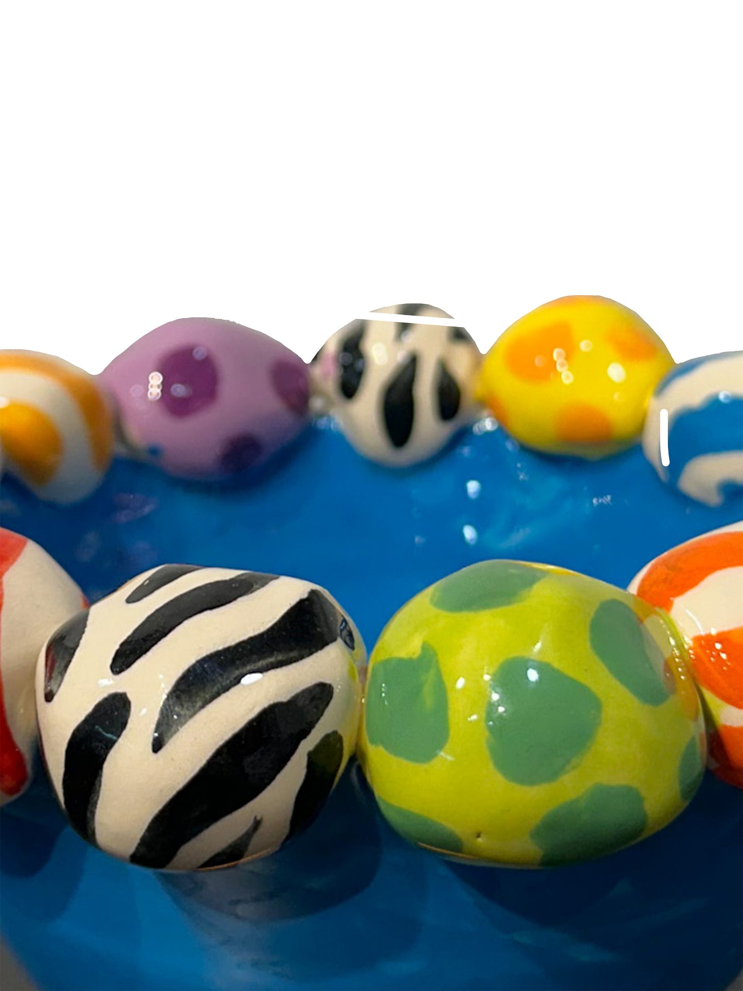 This handmade ceramic footed bowl by Mary Rose Young features a vivid blue glaze and playful hand-painted orbs in stripes, polka dots, and animal prints around its rim. It rests on sculpted red, green, and yellow feet, making it a striking decorative piece suitable for serving or display. Dimensions are approximately 6.5 inches in width and depth, and 4 inches high. Crafted from ceramic, this unique item combines functional art with bold, colorful design.