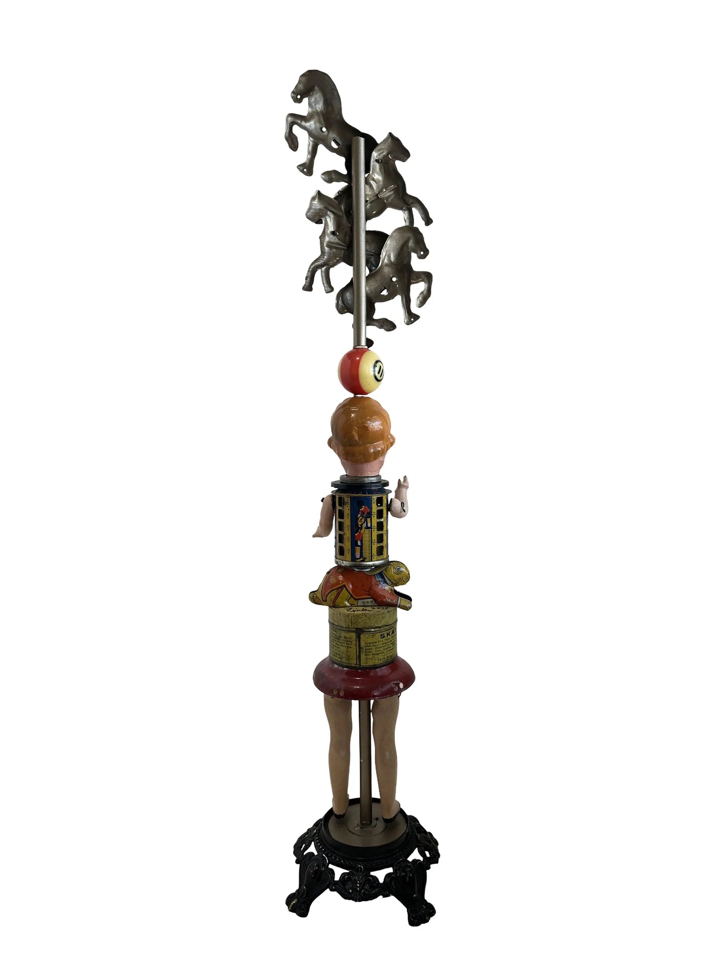 This sculpture, titled 'Skat Horses' by William Fink, is a whimsical standing piece made from vintage and antique components. It features a papier-mâché doll head with antique toy horses on top, a vintage Skat soap tin, a 1940s savings bank, a toy rabbit, and a spinning metal top, all arranged to evoke nostalgia and imagination. Measuring approximately 8.5 inches wide, 39 inches tall, and 8.5 inches deep, this mixed media artwork celebrates creativity through its playful use of found objects.