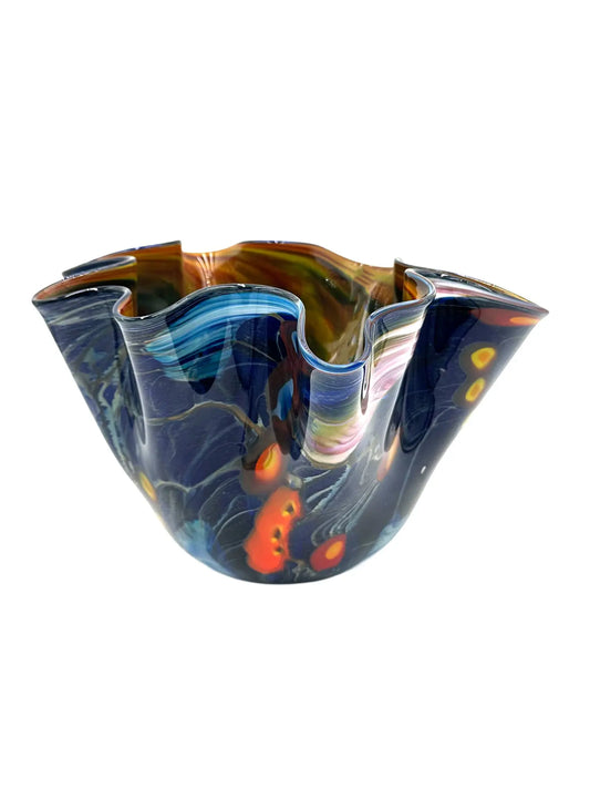 Galaxy Bowl by Steve Palmer is an elegant, handblown glass vase featuring swirling deep blue, silver, orange, and red patterns reminiscent of a cosmic dance. With a fluted rim, it combines artistry and function, ideal for striking decor or floral displays.