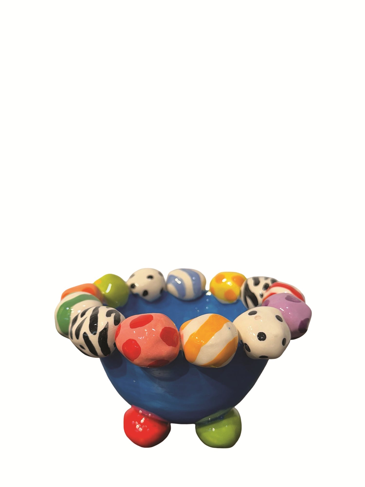 This handmade ceramic footed bowl by Mary Rose Young features a vivid blue glaze and playful hand-painted orbs in stripes, polka dots, and animal prints around its rim. It rests on sculpted red, green, and yellow feet, making it a striking decorative piece suitable for serving or display. Dimensions are approximately 6.5 inches in width and depth, and 4 inches high. Crafted from ceramic, this unique item combines functional art with bold, colorful design.