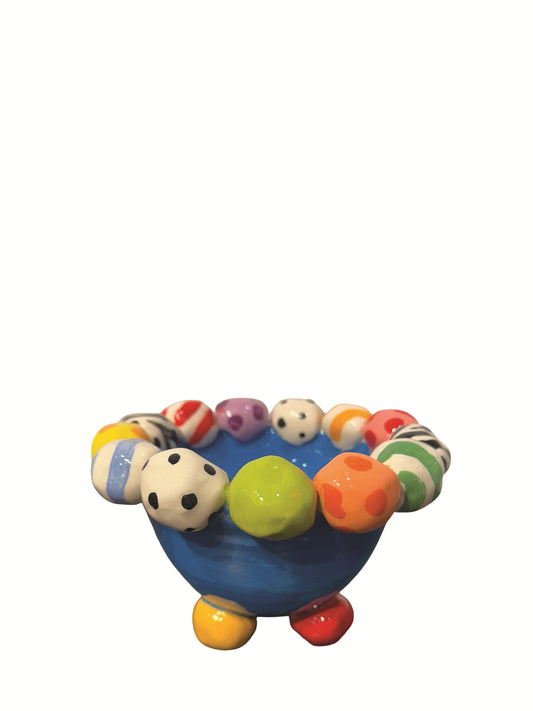 This handmade ceramic footed bowl by Mary Rose Young features a vivid blue glaze and playful hand-painted orbs in stripes, polka dots, and animal prints around its rim. It rests on sculpted red, green, and yellow feet, making it a striking decorative piece suitable for serving or display. Dimensions are approximately 6.5 inches in width and depth, and 4 inches high. Crafted from ceramic, this unique item combines functional art with bold, colorful design.