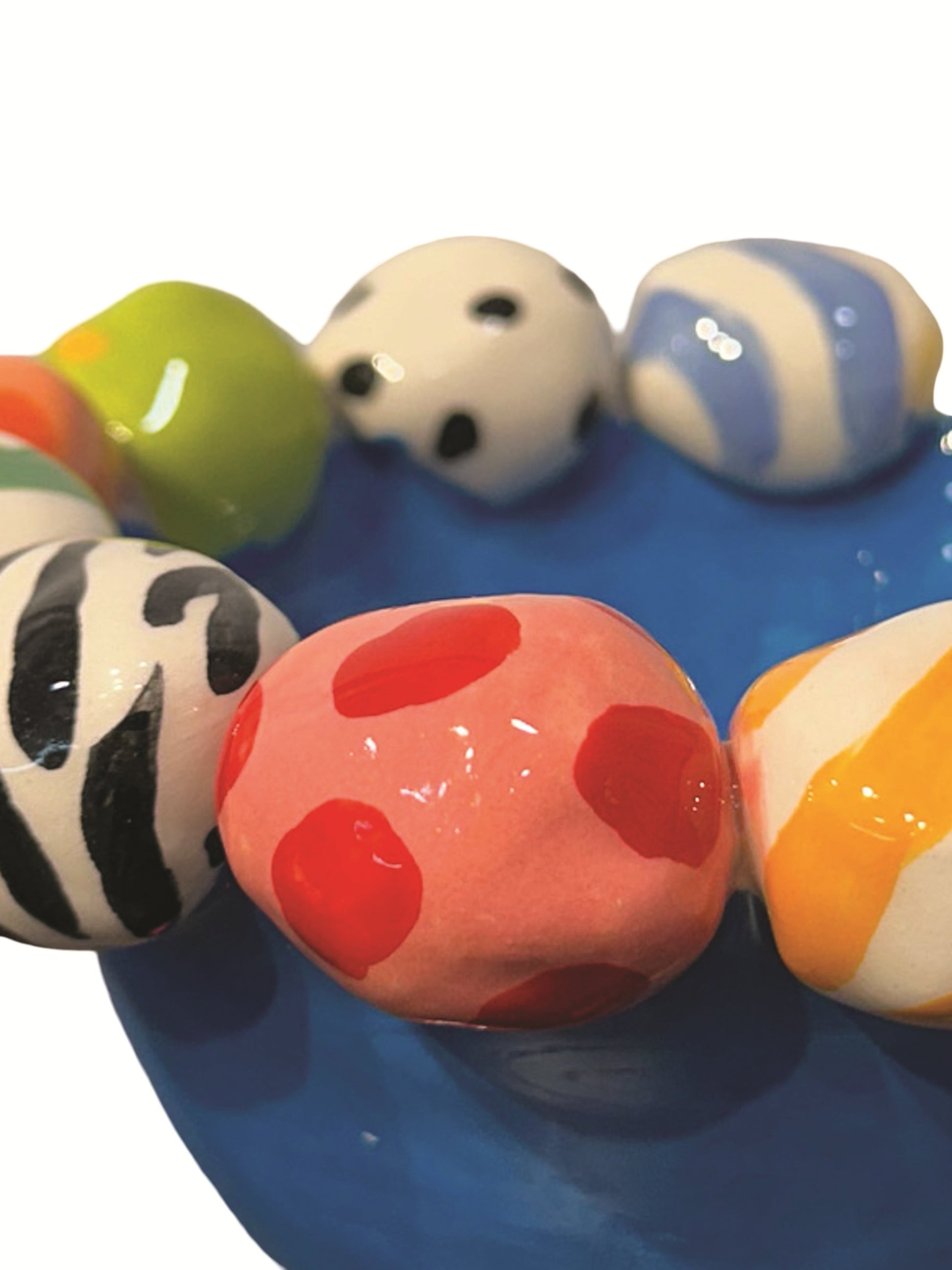 This handmade ceramic footed bowl by Mary Rose Young features a vivid blue glaze and playful hand-painted orbs in stripes, polka dots, and animal prints around its rim. It rests on sculpted red, green, and yellow feet, making it a striking decorative piece suitable for serving or display. Dimensions are approximately 6.5 inches in width and depth, and 4 inches high. Crafted from ceramic, this unique item combines functional art with bold, colorful design.