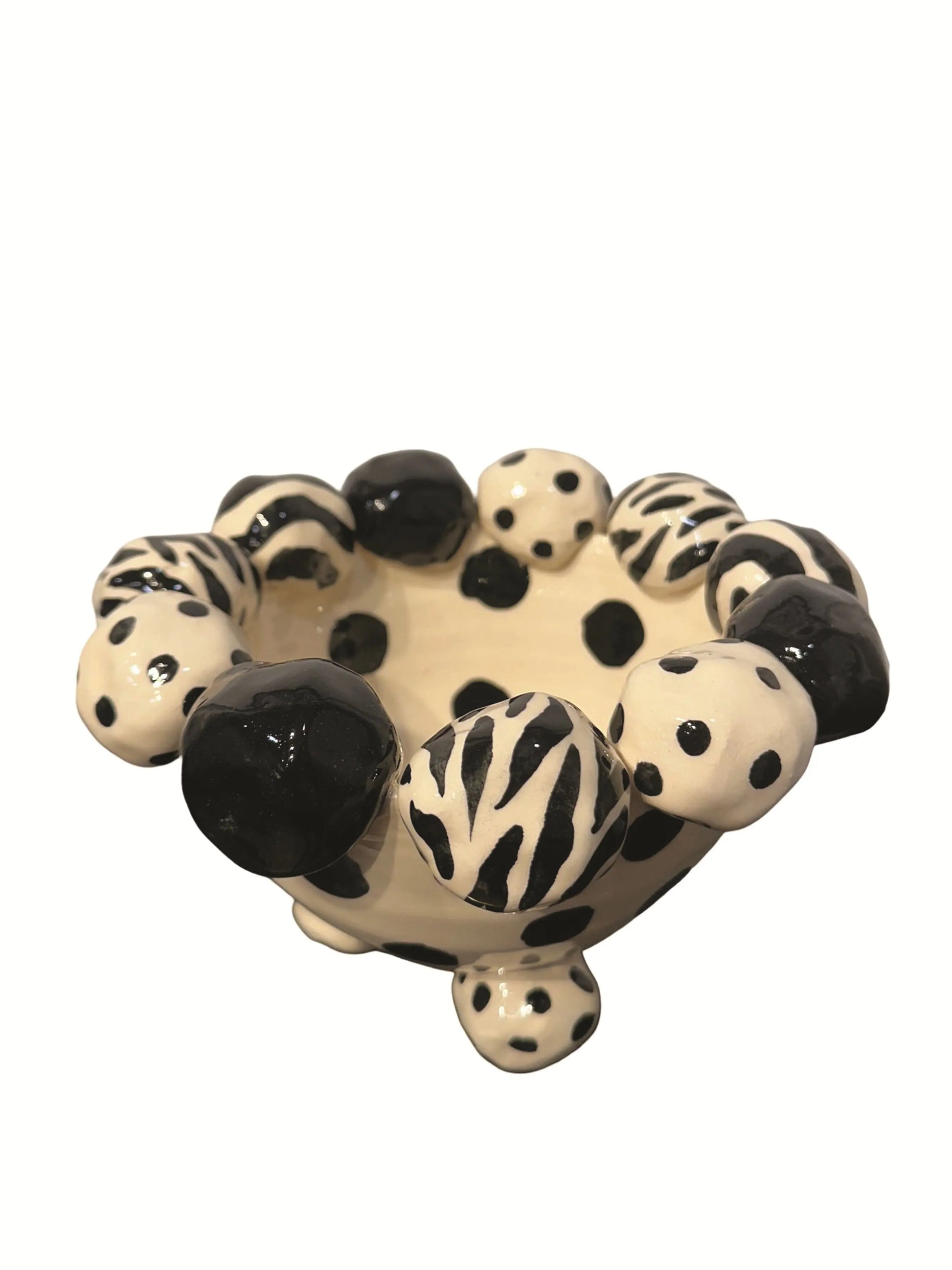 This handmade ceramic footed bowl, designed by Mary Rose Young, features a bold black and cream pattern with hand-painted dots, zebra stripes, and solid black accents. The sculpted dotted feet complement the playful rim, adding charm and balance. Measuring approximately 8 inches wide, 8 inches deep, and 5 inches high, this striking piece serves as both a functional bowl and a decorative statement, perfect for enhancing any table, shelf, or display with artistic flair.