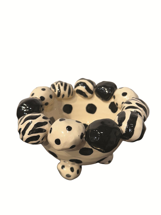 This handmade ceramic footed bowl, designed by Mary Rose Young, features a bold black and cream pattern with hand-painted dots, zebra stripes, and solid black accents. The sculpted dotted feet complement the playful rim, adding charm and balance. Measuring approximately 8 inches wide, 8 inches deep, and 5 inches high, this striking piece serves as both a functional bowl and a decorative statement, perfect for enhancing any table, shelf, or display with artistic flair.