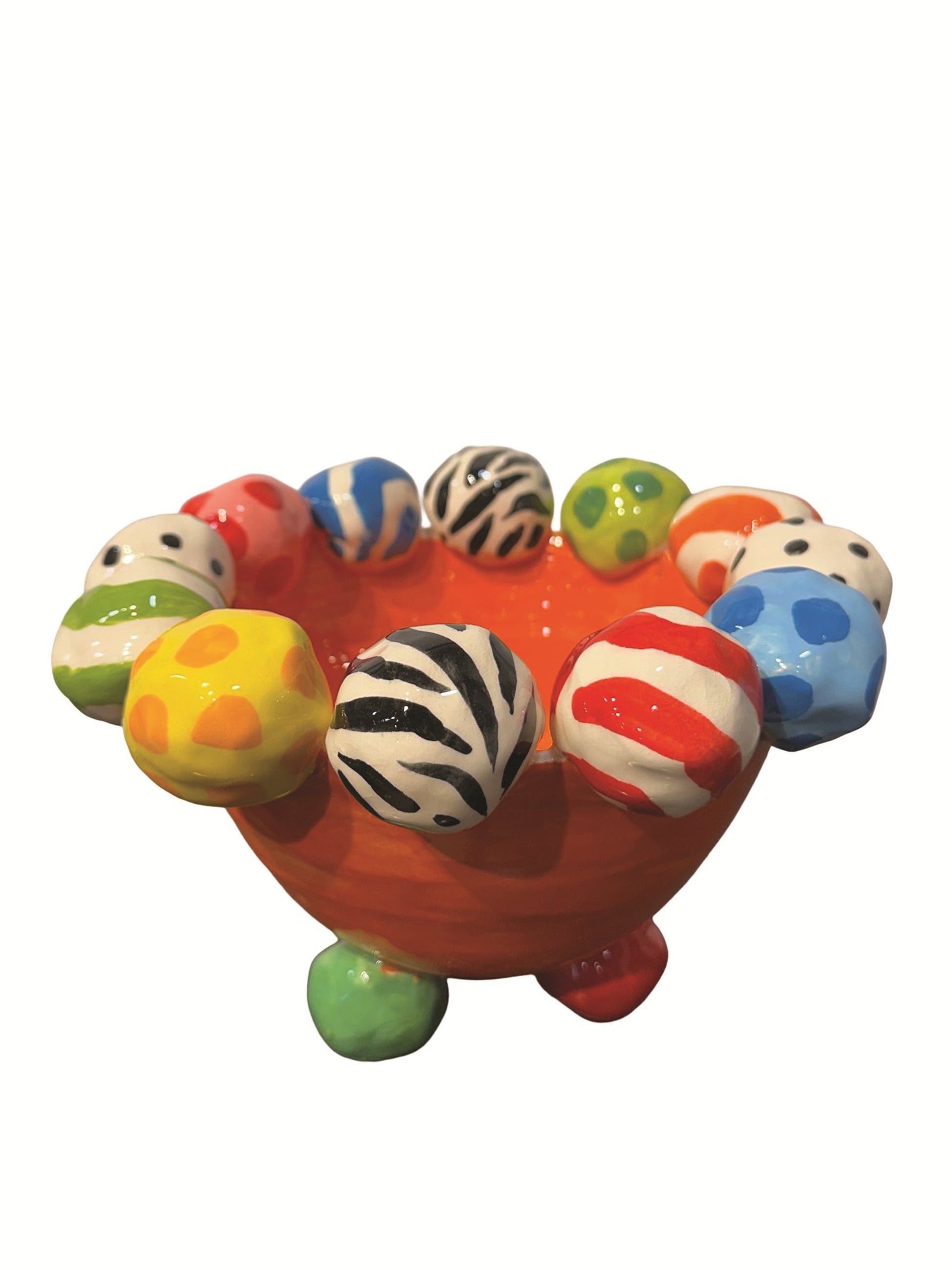 This vibrant handmade ceramic footed bowl by artist Mary Rose Young features a bold orange base with a rim decorated with hand-painted orbs in patterns like zebra stripes, polka dots, and abstract swirls, all in lively colors. Resting on sculpted red, green, and yellow feet, this large, striking piece measures approximately 8.5 inches in width and depth and 5.5 inches in height. It is perfect for both serving and decorative purposes. 