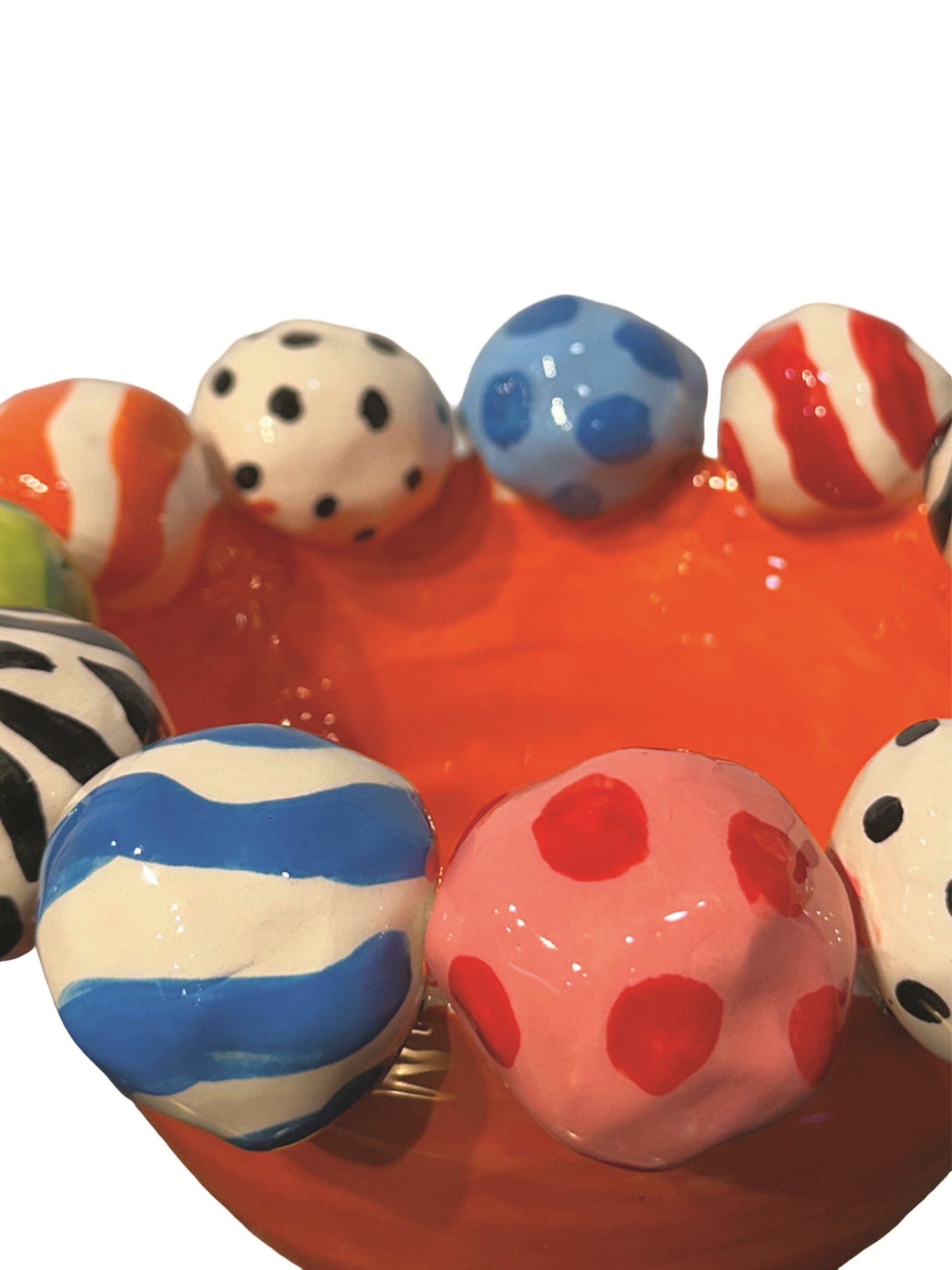 This vibrant handmade ceramic footed bowl by artist Mary Rose Young features a bold orange base with a rim decorated with hand-painted orbs in patterns like zebra stripes, polka dots, and abstract swirls, all in lively colors. Resting on sculpted red, green, and yellow feet, this large, striking piece measures approximately 8.5 inches in width and depth and 5.5 inches in height. It is perfect for both serving and decorative purposes. 