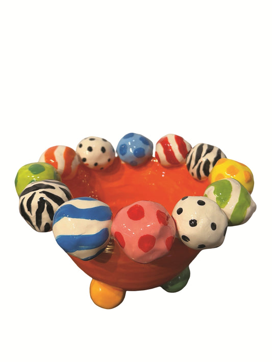 This vibrant handmade ceramic footed bowl by artist Mary Rose Young features a bold orange base with a rim decorated with hand-painted orbs in patterns like zebra stripes, polka dots, and abstract swirls, all in lively colors. Resting on sculpted red, green, and yellow feet, this large, striking piece measures approximately 8.5 inches in width and depth and 5.5 inches in height. It is perfect for both serving and decorative purposes. 