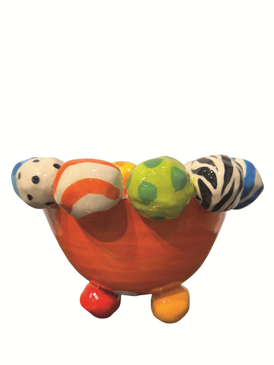 This vibrant handmade ceramic footed bowl by artist Mary Rose Young features a bold orange base with a rim decorated with hand-painted orbs in patterns like zebra stripes, polka dots, and abstract swirls, all in lively colors. Resting on sculpted red, green, and yellow feet, this large, striking piece measures approximately 8.5 inches in width and depth and 5.5 inches in height. It is perfect for both serving and decorative purposes. 