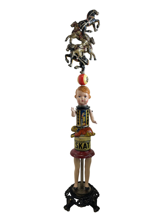 This sculpture, titled 'Skat Horses' by William Fink, is a whimsical standing piece made from vintage and antique components. It features a papier-mâché doll head with antique toy horses on top, a vintage Skat soap tin, a 1940s savings bank, a toy rabbit, and a spinning metal top, all arranged to evoke nostalgia and imagination. Measuring approximately 8.5 inches wide, 39 inches tall, and 8.5 inches deep, this mixed media artwork celebrates creativity through its playful use of found objects.