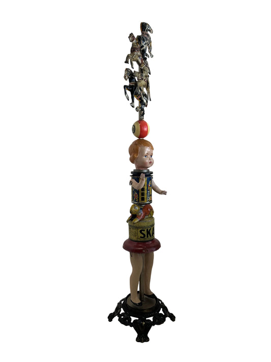 This sculpture, titled 'Skat Horses' by William Fink, is a whimsical standing piece made from vintage and antique components. It features a papier-mâché doll head with antique toy horses on top, a vintage Skat soap tin, a 1940s savings bank, a toy rabbit, and a spinning metal top, all arranged to evoke nostalgia and imagination. Measuring approximately 8.5 inches wide, 39 inches tall, and 8.5 inches deep, this mixed media artwork celebrates creativity through its playful use of found objects.
