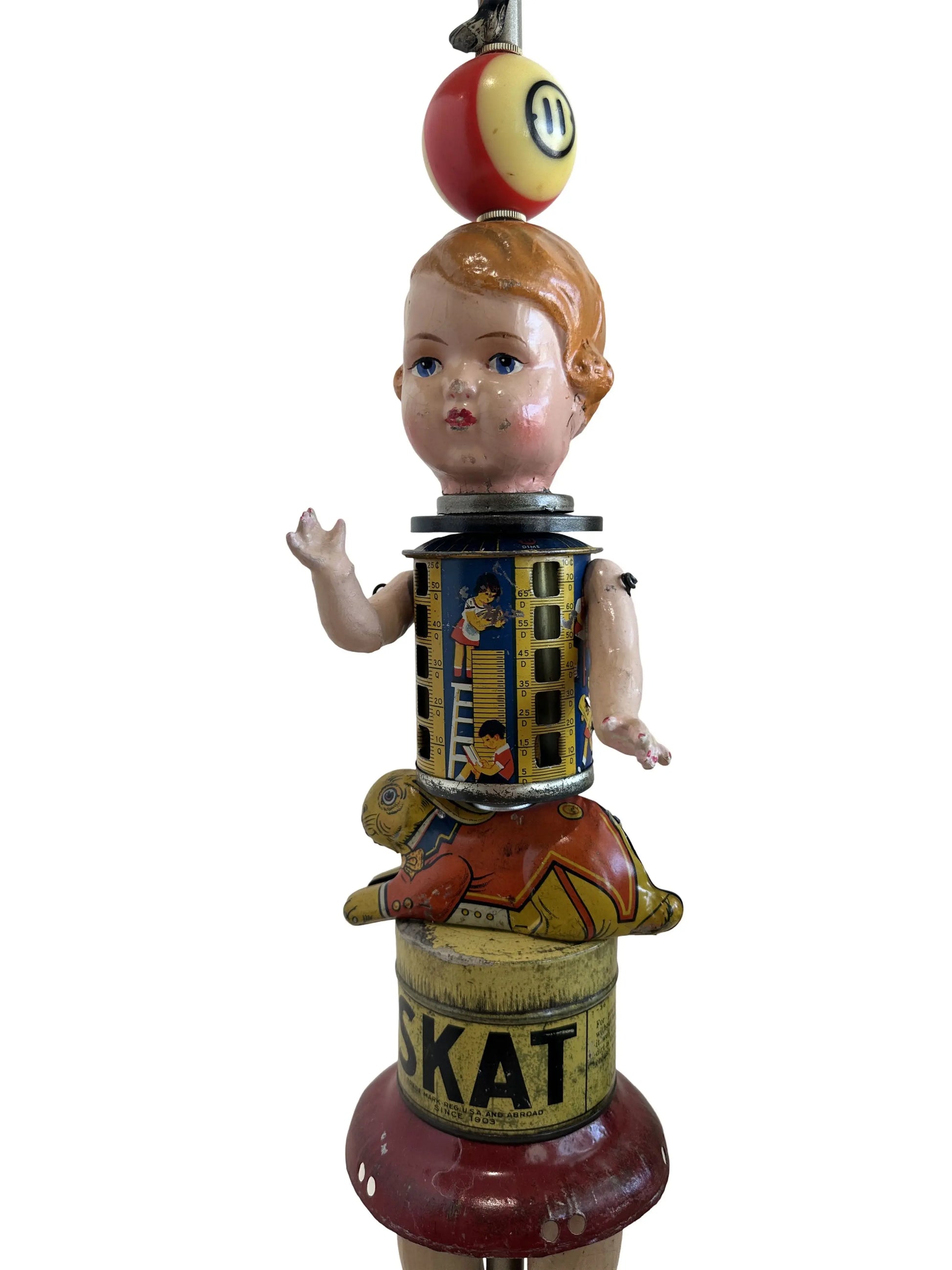 This sculpture, titled 'Skat Horses' by William Fink, is a whimsical standing piece made from vintage and antique components. It features a papier-mâché doll head with antique toy horses on top, a vintage Skat soap tin, a 1940s savings bank, a toy rabbit, and a spinning metal top, all arranged to evoke nostalgia and imagination. Measuring approximately 8.5 inches wide, 39 inches tall, and 8.5 inches deep, this mixed media artwork celebrates creativity through its playful use of found objects.