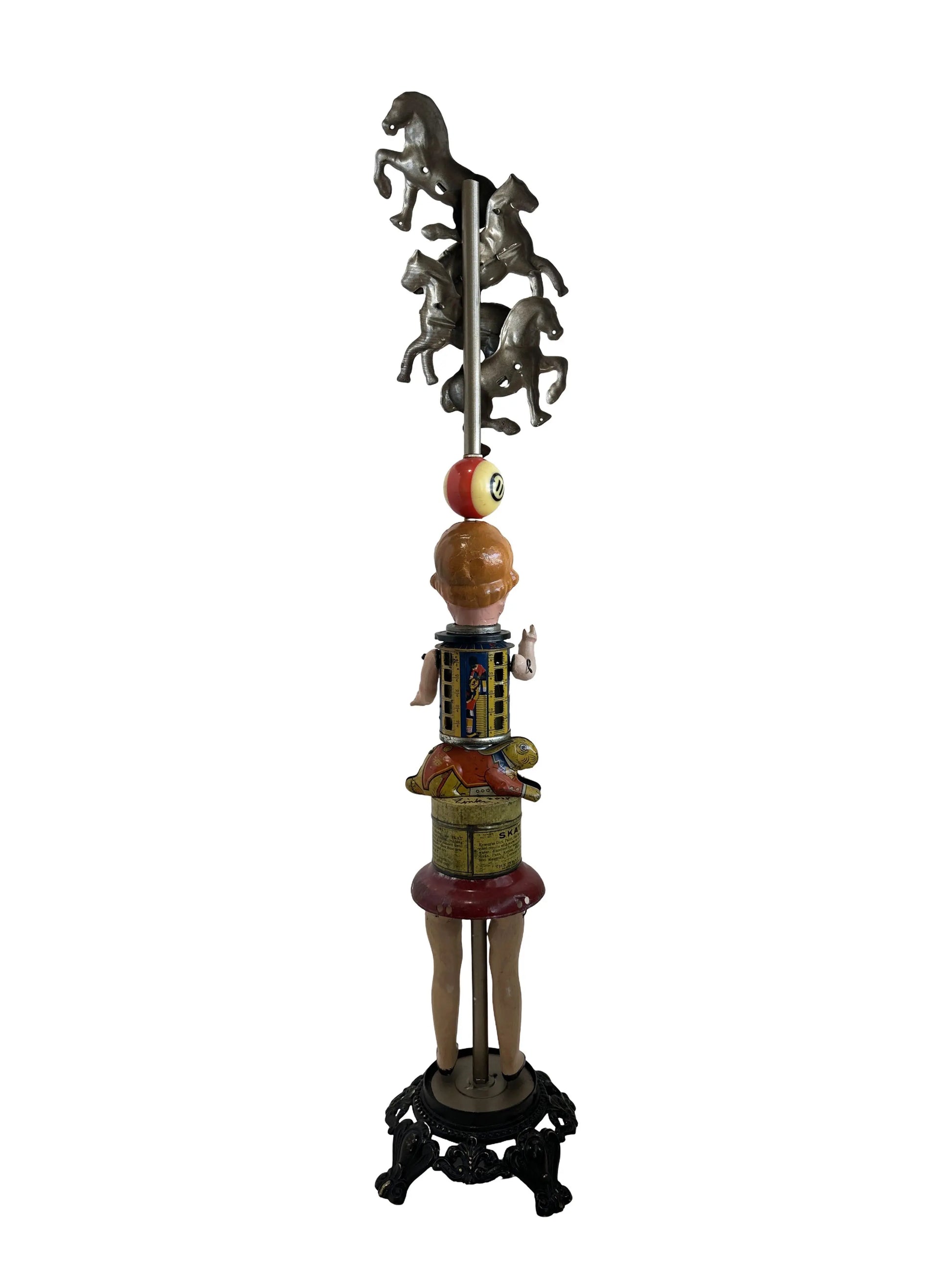 This sculpture, titled 'Skat Horses' by William Fink, is a whimsical standing piece made from vintage and antique components. It features a papier-mâché doll head with antique toy horses on top, a vintage Skat soap tin, a 1940s savings bank, a toy rabbit, and a spinning metal top, all arranged to evoke nostalgia and imagination. Measuring approximately 8.5 inches wide, 39 inches tall, and 8.5 inches deep, this mixed media artwork celebrates creativity through its playful use of found objects.