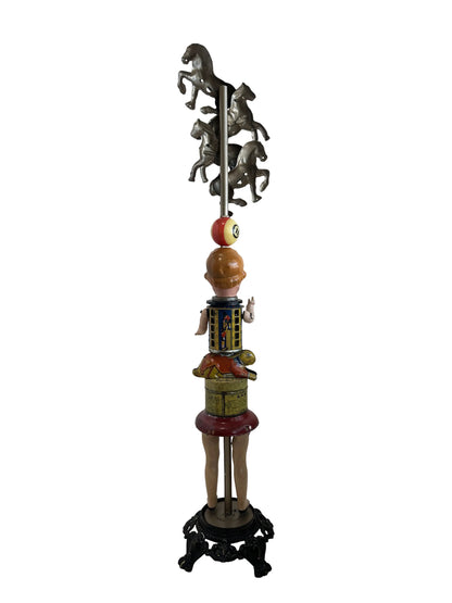 This sculpture, titled 'Skat Horses' by William Fink, is a whimsical standing piece made from vintage and antique components. It features a papier-mâché doll head with antique toy horses on top, a vintage Skat soap tin, a 1940s savings bank, a toy rabbit, and a spinning metal top, all arranged to evoke nostalgia and imagination. Measuring approximately 8.5 inches wide, 39 inches tall, and 8.5 inches deep, this mixed media artwork celebrates creativity through its playful use of found objects.