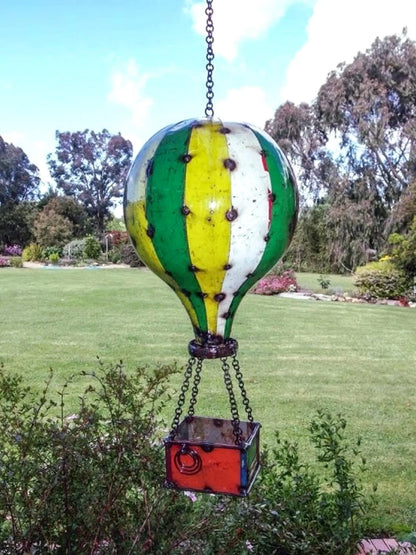 A vintage-style hot air balloon garden swing hanging from a chain in a backyard garden. The balloon features vertical stripes of green, yellow, and white with rusted details, and it hangs above a small red and green basket.