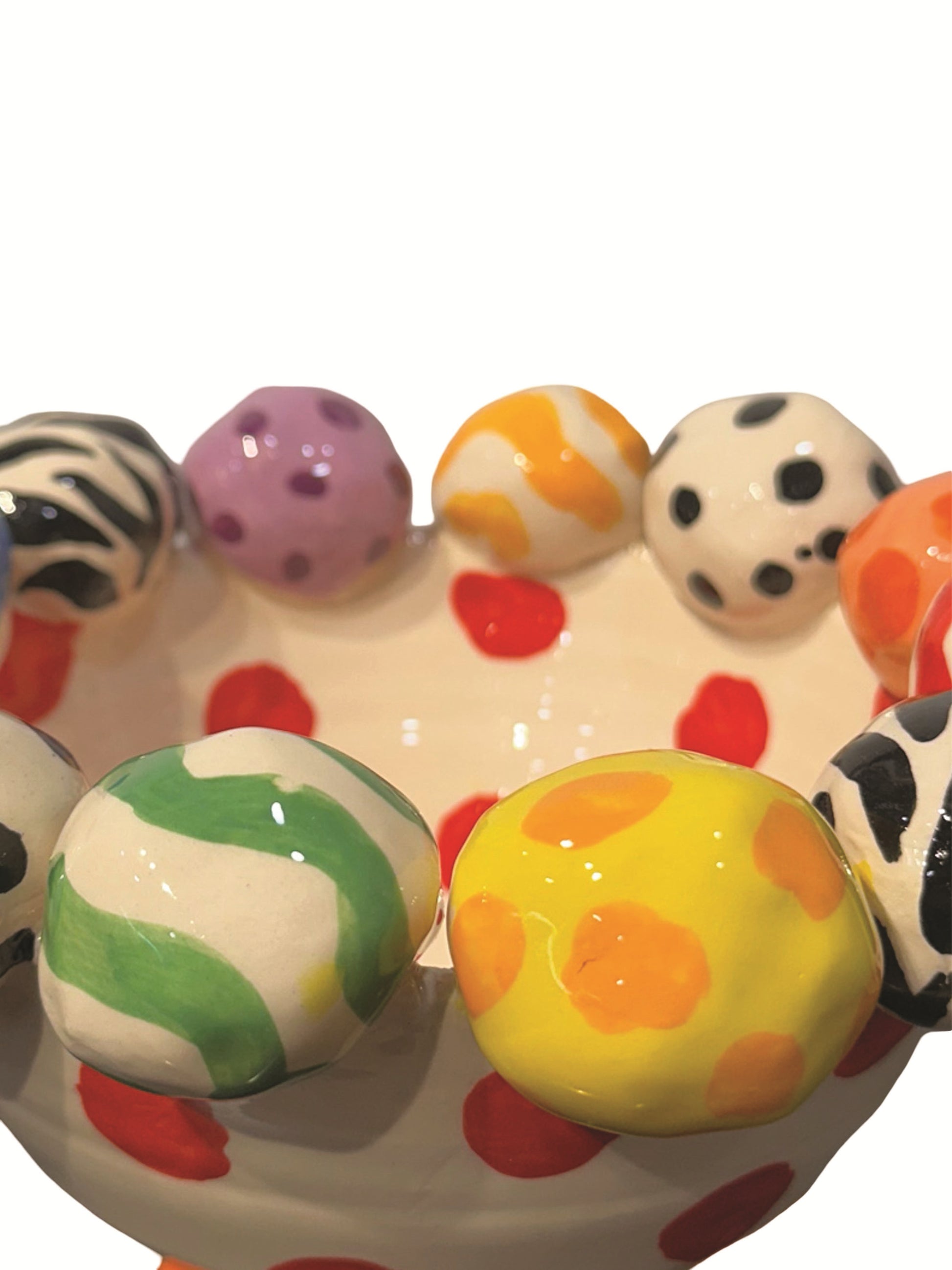 This handmade ceramic-footed bowl by Mary Rose Young features a whimsical design with a creamy white base decorated with bold red polka dots. The rim is adorned with hand-painted orbs displaying various patterns such as zebra stripes, colorful swirls, and polka dots. Resting on sculpted orange, blue, and gold feet, this vibrant piece is ideal for serving or decorative use, measuring approximately 8.5 inches in width and depth and 5.5 inches tall.