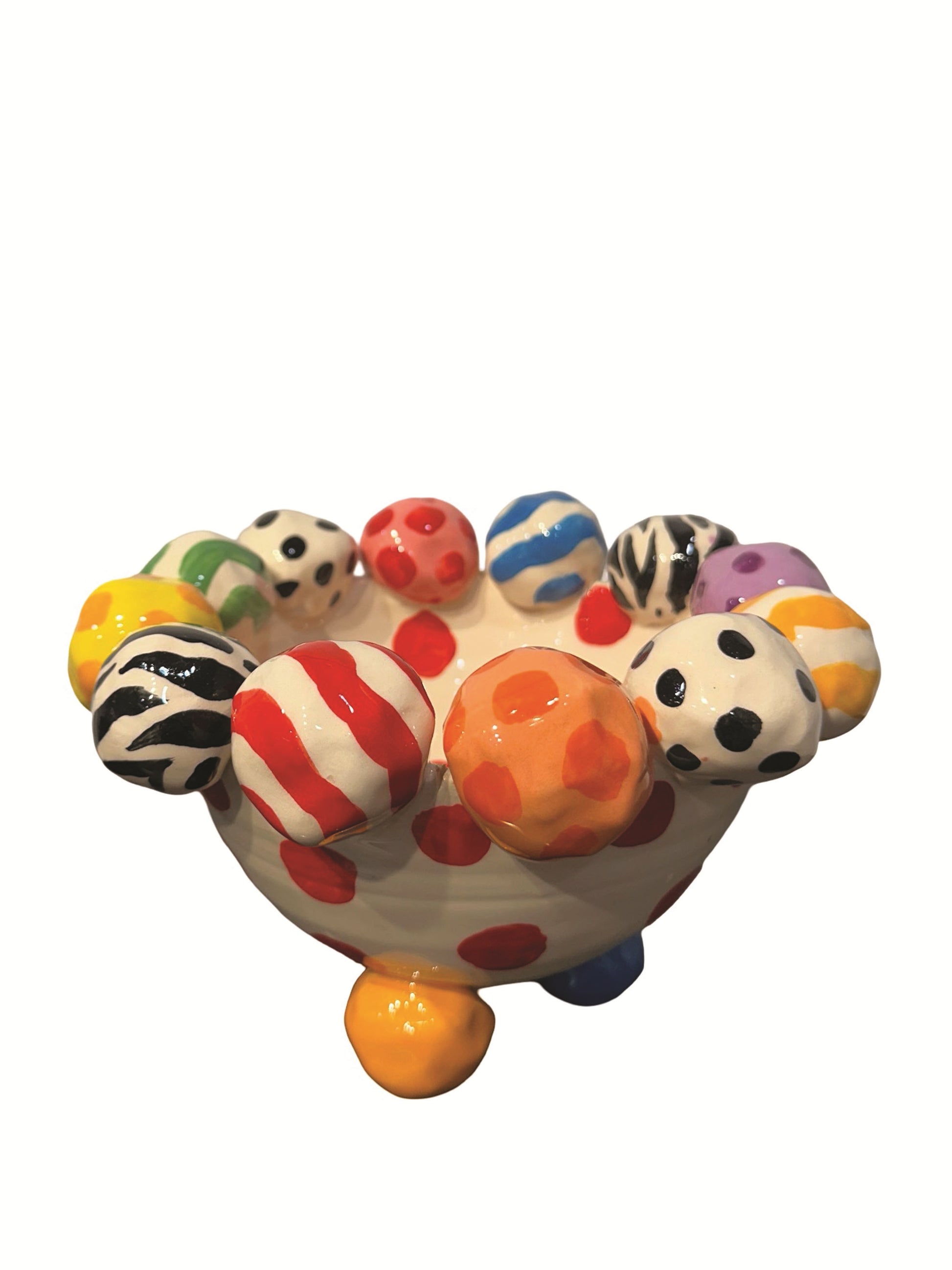 This handmade ceramic-footed bowl by Mary Rose Young features a whimsical design with a creamy white base decorated with bold red polka dots. The rim is adorned with hand-painted orbs displaying various patterns such as zebra stripes, colorful swirls, and polka dots. Resting on sculpted orange, blue, and gold feet, this vibrant piece is ideal for serving or decorative use, measuring approximately 8.5 inches in width and depth and 5.5 inches tall.