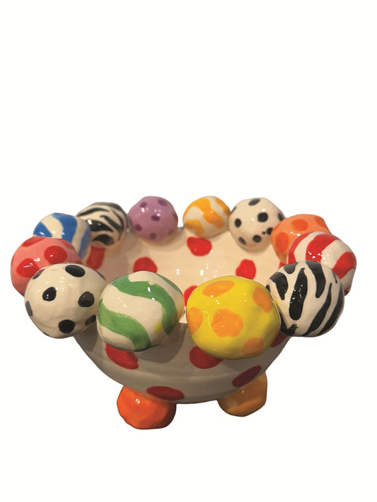 This handmade ceramic-footed bowl by Mary Rose Young features a whimsical design with a creamy white base decorated with bold red polka dots. The rim is adorned with hand-painted orbs displaying various patterns such as zebra stripes, colorful swirls, and polka dots. Resting on sculpted orange, blue, and gold feet, this vibrant piece is ideal for serving or decorative use, measuring approximately 8.5 inches in width and depth and 5.5 inches tall.